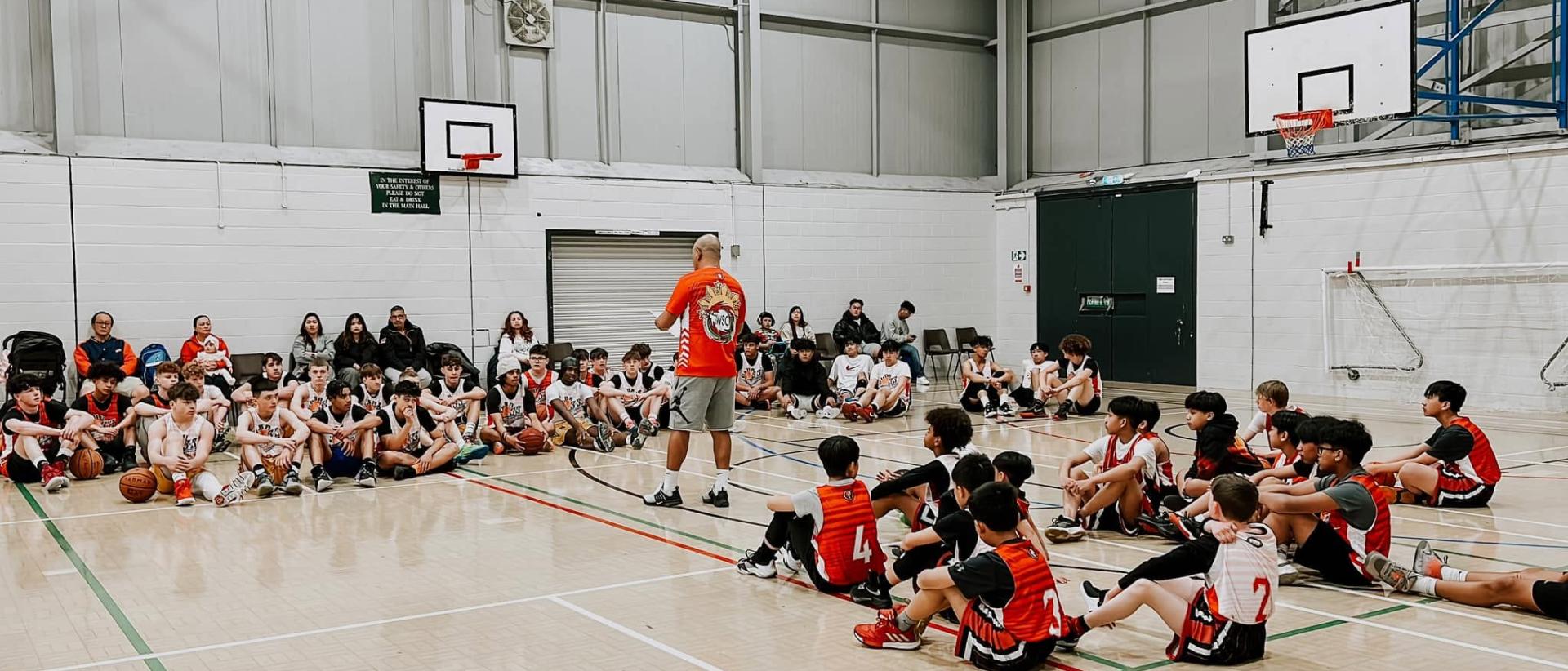 coach leo talking to his players sat on the floor of a basketball gym
